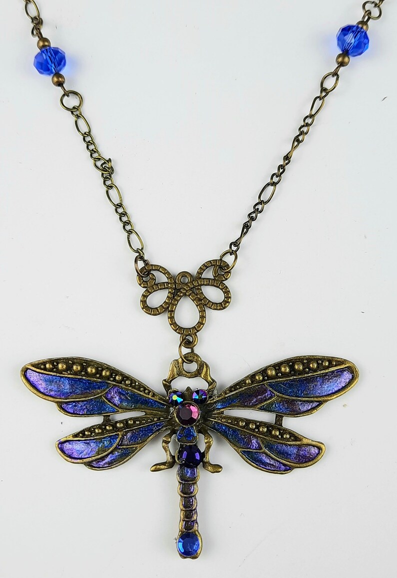 May include: A bronze necklace with a blue dragonfly pendant. The dragonfly has iridescent wings and a purple rhinestone body. The necklace has a chain with blue crystal beads.