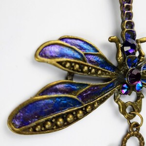 May include: A gold dragonfly pendant with iridescent blue and purple wings and purple rhinestones. The dragonfly is hanging from a gold chain with a circular charm.