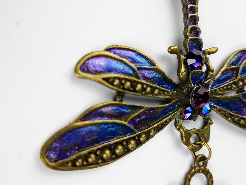 May include: A gold dragonfly pendant with iridescent blue and purple wings and purple rhinestones. The dragonfly is hanging from a gold chain with a circular charm.