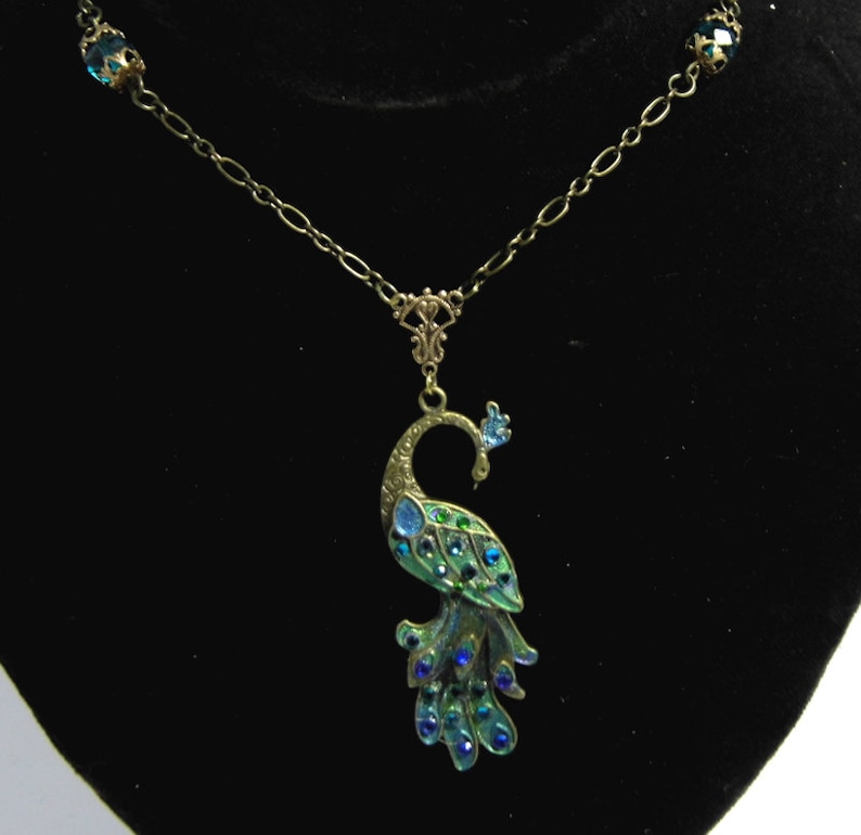 May include: A gold chain necklace with a peacock pendant. The peacock is made of metal and has blue and green enamel with rhinestones. The chain has two blue rhinestone accents.