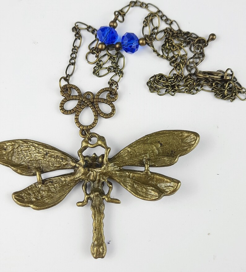 May include: A gold-toned dragonfly pendant necklace with blue glass beads. The dragonfly has intricate details and is attached to a delicate chain.