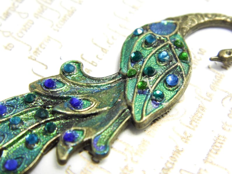 May include: A peacock-shaped metal charm with green and blue enamel and rhinestones. The charm is in a vintage style and has a detailed design.