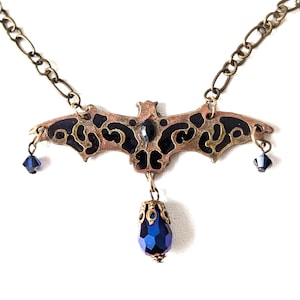 May include: A black and gold bat pendant necklace with blue crystal accents. The bat is detailed with intricate patterns and has a dangling blue crystal drop.