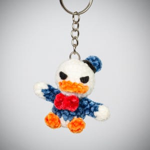 Peut inclure: Porte-clés fait à la main représentant le personnage de Donald Duck en peluche. Le porte-clés a une tête blanche, un bec orange et une tenue bleue et blanche avec un nœud papillon rouge. Le porte-clés est attaché à un anneau en métal argenté.