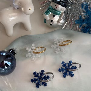 Peut inclure: Une composition festive avec des décorations de Noël et des ronds de serviette en forme de flocons de neige. Les décorations comprennent un renne blanc avec des accents dorés, un bonhomme de neige et une sphère bleue. Les ronds de serviette sont transparents et dorés, et bleus et argentés.