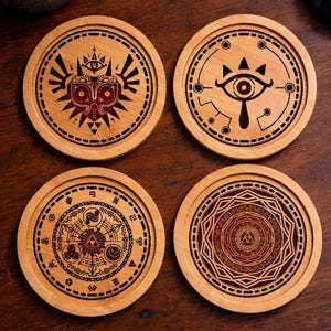 May include: A set of four circular light brown wooden coasters, each featuring a distinct dark brown laser-engraved design. The designs include a mask, an eye symbol, a complex circular emblem with geometric patterns, and a detailed mandala-like motif.