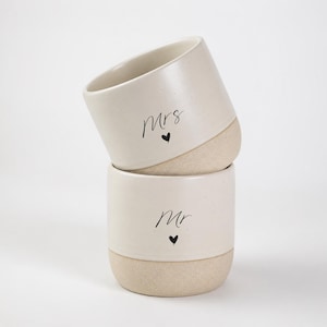 Puede incluir: Dos tazas de cerámica apiladas con un esmalte crema moteado en la parte superior y una base beige rugosa y texturizada. La taza superior presenta el texto negro "Mrs" con un pequeño corazón, y la inferior el texto "mr" con un corazón.