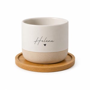 Może przedstawiać: Cylindryczny ceramiczny kubek z matową białą górną częścią i naturalną beżową podstawą. Na kubku widnieje imię Helena wydrukowane czarną czcionką pisankową nad małą czarną ikoną serca. Kubek stoi na okrągłej podstawce wykonanej z drewna bambusowego.