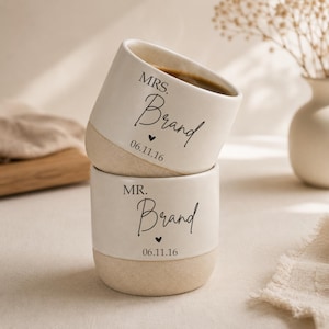 Può includere: Due tazze in ceramica impilate con un design bicolore caratterizzato da una parte superiore bianca liscia e una base beige testurizzata. La tazza superiore riporta la scritta MRS. Brand e 06.11.16, quella inferiore MR. Brand e 06.11.16.