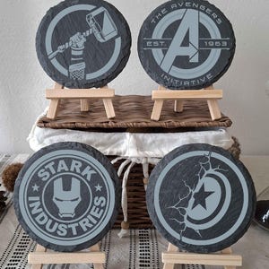 Può includere: Quattro sottobicchieri rotondi in ardesia con disegni a tema supereroi. I sottobicchieri presentano il logo degli Avengers, il martello di Thor, il logo Stark Industries e lo scudo di Capitan America. Ogni sottobicchiere è esposto su un piccolo cavalletto di legno.