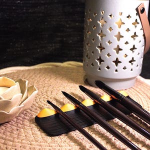 Peut inclure: Un photophore en céramique blanche avec des découpes en forme d'étoiles et une poignée en cuir marron. L'image montre également un ensemble de pinceaux et un bougeoir décoratif en forme de fleur, le tout disposé sur une surface texturée.