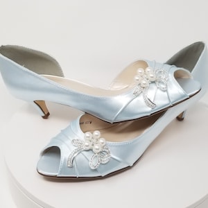 A pair of baby blue satin kitten heel shoes with a peep toe and a pearl and crystal bow design on the side of the shoes