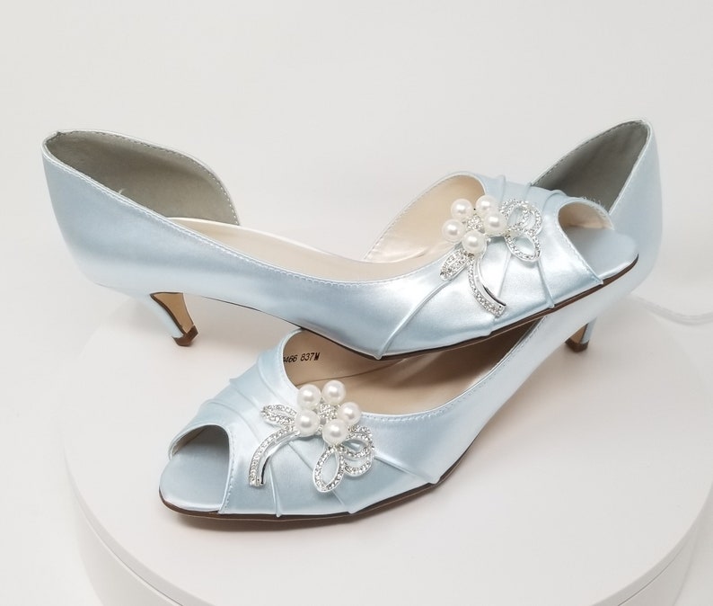 A pair of baby blue satin kitten heel shoes with a peep toe and a pearl and crystal bow design on the side of the shoes