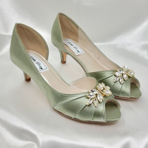 a pair of sage green satin kitten heel shoes with a peep toe and a gold and crystal design on the front of the shoes