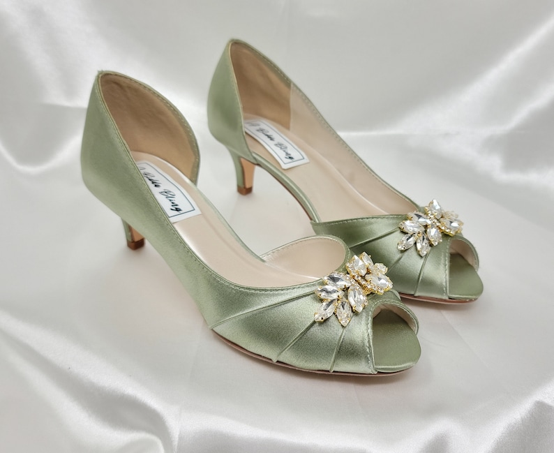 a pair of sage green satin kitten heel shoes with a peep toe and a gold and crystal design on the front of the shoes