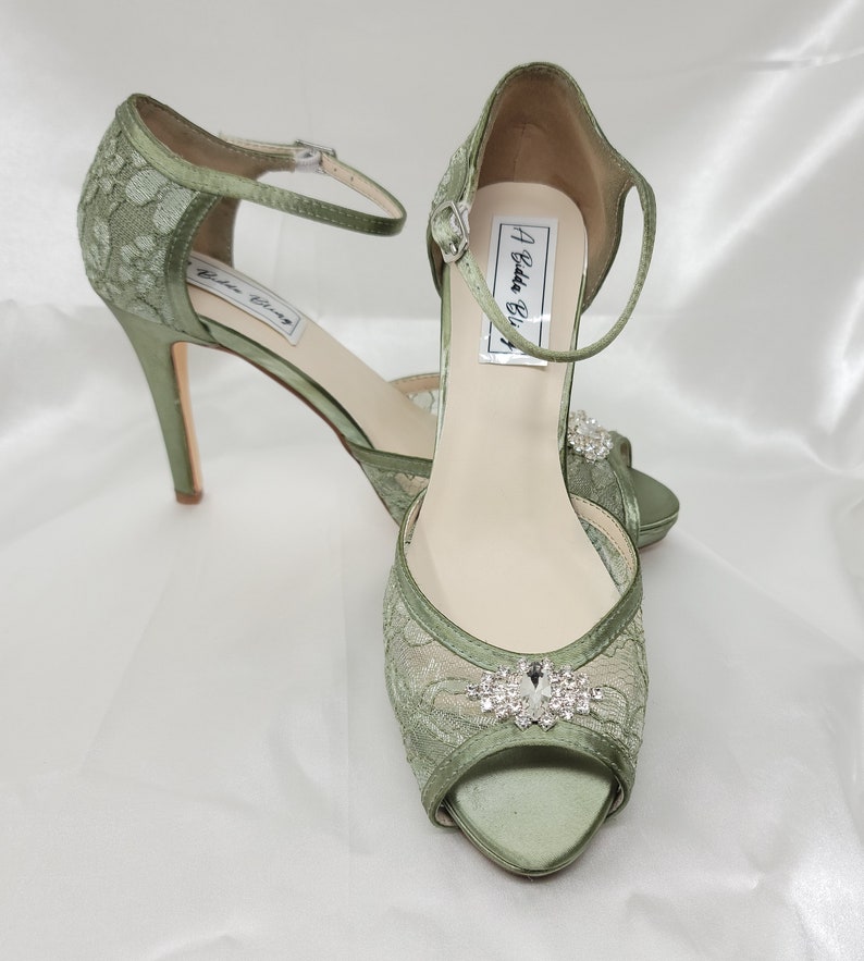 A pair of sage green high heeled platform lace shoes with an ankle strap and a crystal design on the front of the peep toe shoe