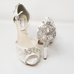 May include: White satin bridal shoes with a lace design and crystal embellishments. The shoes have an ankle strap and a high heel.