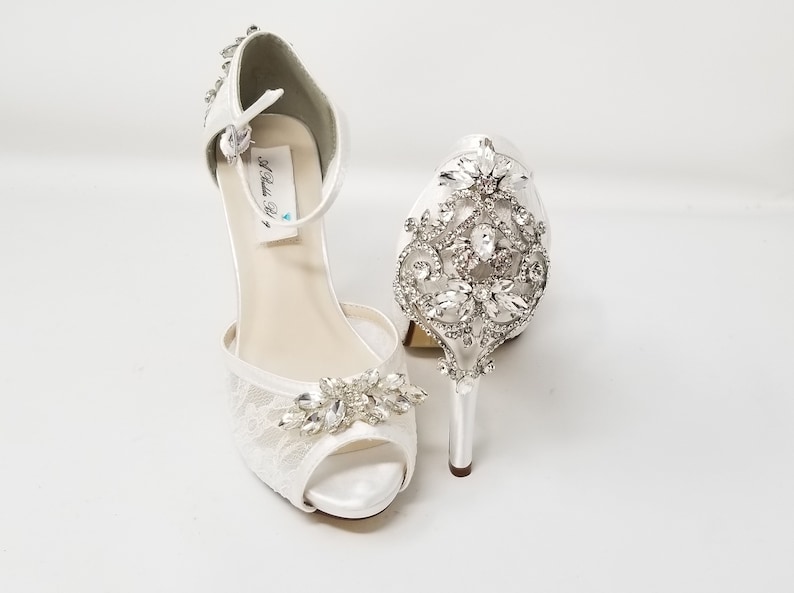 May include: White satin bridal shoes with a lace design and crystal embellishments. The shoes have an ankle strap and a high heel.