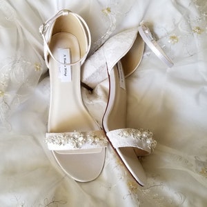 A pair of ivory lace covered low block heel shoes with an ankle strap and a crystal and pearl design on the front toe strap of the shoes