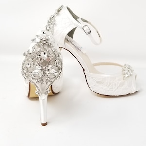 May include: White satin bridal shoes with a peep toe and ankle strap. The shoes feature a large rhinestone embellishment on the front.