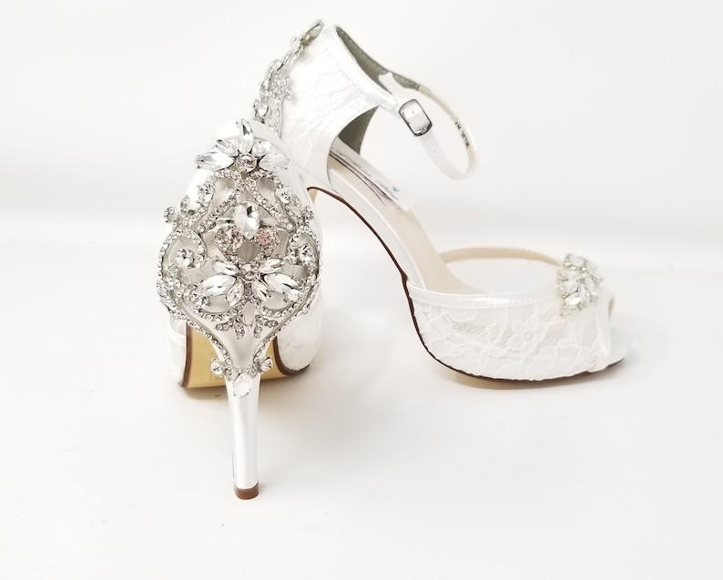 May include: White satin bridal shoes with a peep toe and ankle strap. The shoes feature a large rhinestone embellishment on the front.