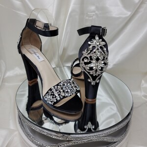 A pair of high black block heel shoes with an ankle strap and a crystal and pearl design on the front toe strap of the shoes and a large crystal design on the back heel of the shoes