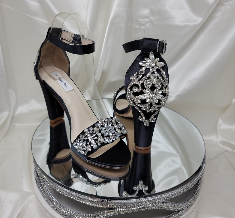 A pair of high black block heel shoes with an ankle strap and a crystal and pearl design on the front toe strap of the shoes and a large crystal design on the back heel of the shoes