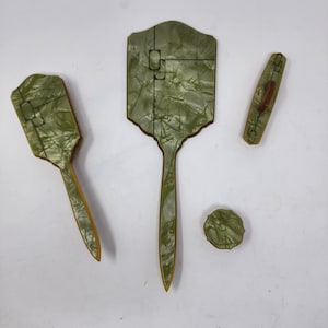 May include: A vintage vanity set in shades of green and brown. The set includes a hand mirror, a hair brush, a clothes brush, and a small round container. The items have a marbled appearance and are arranged on a white surface.
