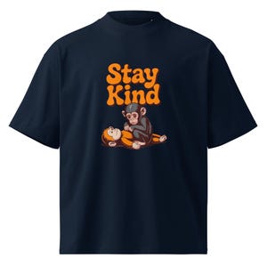 May include: A dark navy blue short-sleeve t-shirt featuring a graphic print of two chimpanzees, one lying down and one sitting on top, with the orange text "Stay Kind" positioned above the monkeys in a bold, rounded, retro-style font.