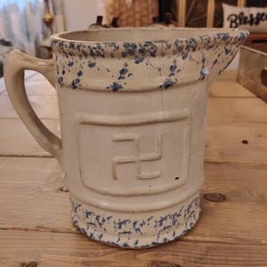 May include: Cream stoneware jug with blue speckles and a handle. The jug features a raised emblem with a swastika design. The rim and base have a blue speckled pattern. The jug is on a wooden surface.