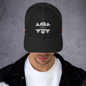 Könnte beinhalten: Schwarze Trucker-Mütze mit weißem, gesticktem Totenkopf- und gekreuzten Knochen-Design. Der Totenkopf trägt einen Hut mit dem Text "Captain 10". Die Mütze hat einen gebogenen Schirm und eine Mesh-Rückseite.