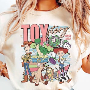 May include: Cream-colored t-shirt featuring a colorful graphic of Toy Story characters, including Woody, Buzz Lightyear, and Rex. The text "TOY STORY" is displayed in pink, with "SINCE 1995" below. A fun, nostalgic design.