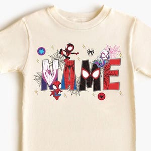 May include: Cream-colored t-shirt featuring the word "NAME" in a colorful, comic-style font. The letters are decorated with Spider-Man characters and spiderwebs. The shirt has short sleeves and a crew neck.