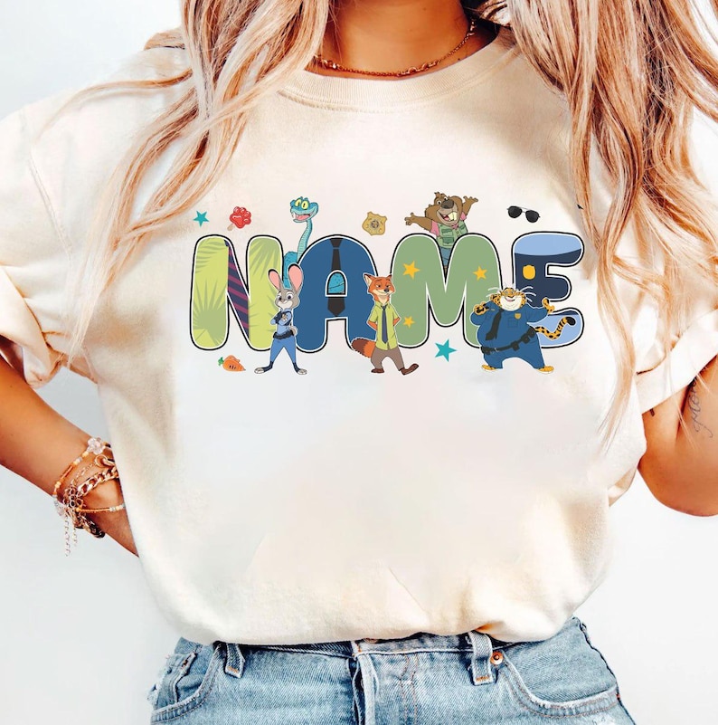 Puede incluir: Camiseta color crema con la palabra "NAME" en una fuente colorida y de dibujos animados con personajes de animales. El dise&ntilde;o incluye un conejo, un zorro, un tigre y otros personajes, con estrellas y detalles florales. Es una prenda de vestir informal.