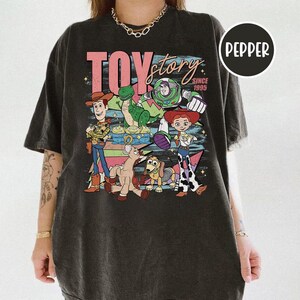 May include: Dark gray t-shirt featuring the words "TOY STORY" in pink and the characters Woody, Buzz Lightyear, Jessie, Rex, and others. The shirt has a vintage, distressed look, with the text "SINCE 1995" below the main title.