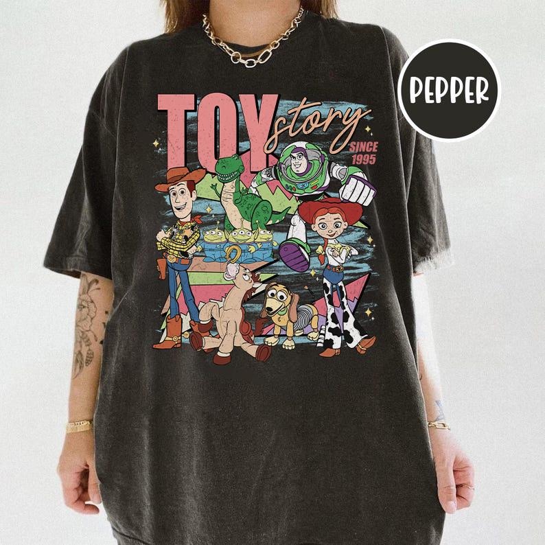 May include: Dark gray t-shirt featuring the words "TOY STORY" in pink and the characters Woody, Buzz Lightyear, Jessie, Rex, and others. The shirt has a vintage, distressed look, with the text "SINCE 1995" below the main title.