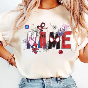 May include: Cream-colored t-shirt featuring the word "NAME" in a colorful, comic-style design. The letters are decorated with spiderwebs and various Spider-Man characters. Small star accents and spider icons complete the design.