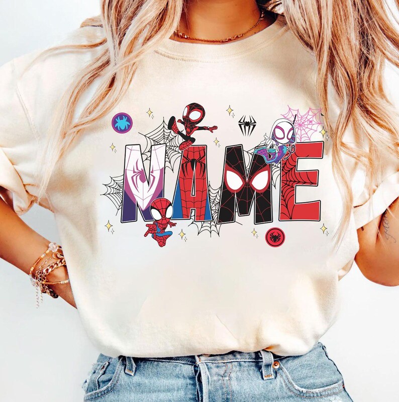 May include: Cream-colored t-shirt featuring the word "NAME" in a colorful, comic-style design. The letters are decorated with spiderwebs and various Spider-Man characters. Small star accents and spider icons complete the design.
