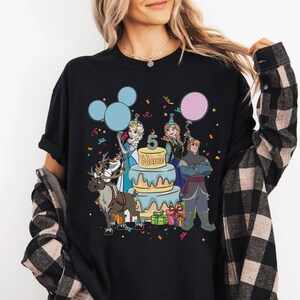 Puede incluir: Camiseta negra con un gr&aacute;fico colorido de personajes de dibujos animados, una tarta de cumplea&ntilde;os con el n&uacute;mero 5 y la palabra "NAME". El dise&ntilde;o incluye globos, confeti y cajas de regalo. Tambi&eacute;n aparece un reno.