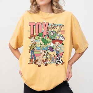 May include: Yellow t-shirt featuring a graphic of Toy Story characters, including Woody, Buzz Lightyear, and Jessie. The text "TOY STORY SINCE 1995" is displayed in a retro font. The shirt has short sleeves and a crew neck.
