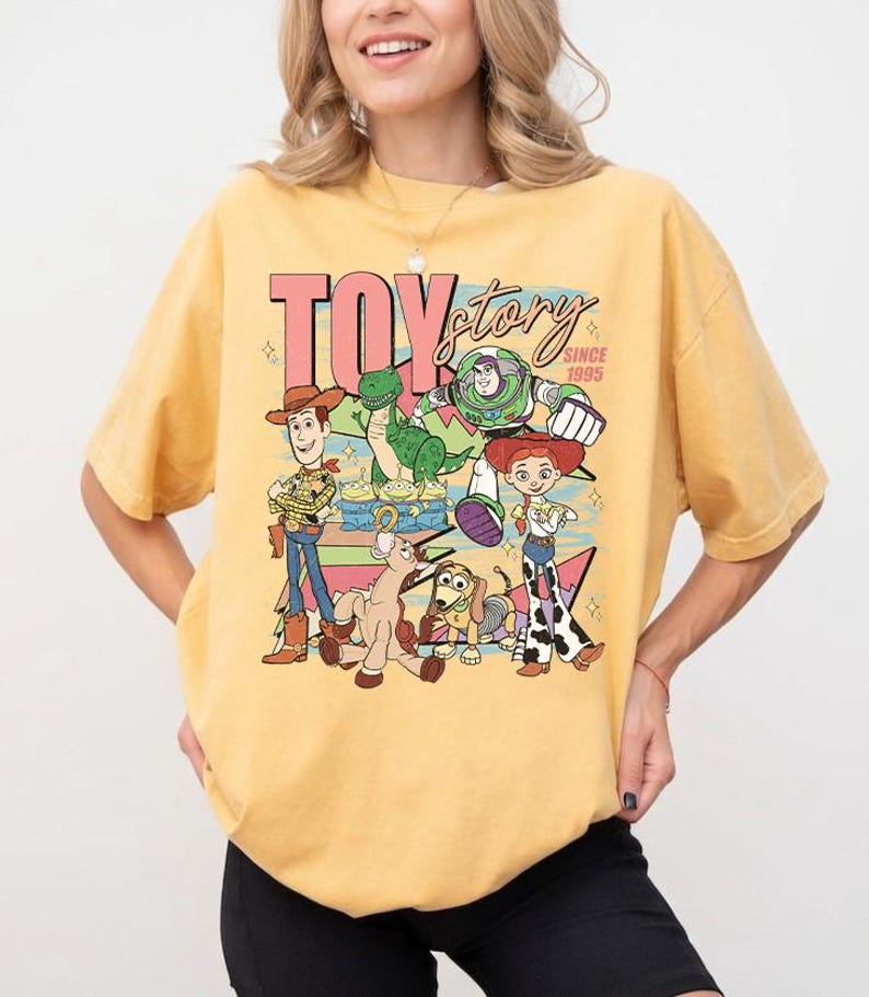 May include: Yellow t-shirt featuring a graphic of Toy Story characters, including Woody, Buzz Lightyear, and Jessie. The text "TOY STORY SINCE 1995" is displayed in a retro font. The shirt has short sleeves and a crew neck.