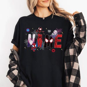 May include: Black t-shirt featuring the word "NAME" in a colorful, comic-style design with spider web accents and various Spider-Man characters. The shirt has a crew neck and short sleeves.
