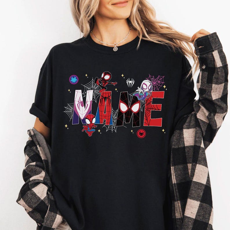 May include: Black t-shirt featuring the word "NAME" in a colorful, comic-style design with spider web accents and various Spider-Man characters. The shirt has a crew neck and short sleeves.