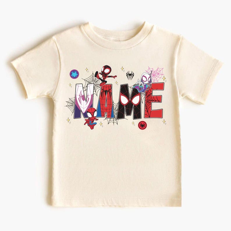 May include: Cream-colored t-shirt featuring the word "NAME" in a colorful, comic-style font. The letters are decorated with Spider-Man characters and spiderwebs. The shirt has short sleeves and a crew neck.