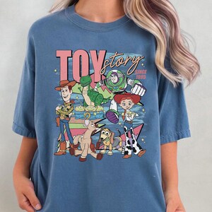 May include: Blue t-shirt featuring a graphic of characters from the animated film "Toy Story." The design includes the text "TOY story" in pink and white, along with various characters like Woody, Buzz Lightyear, and Jessie.