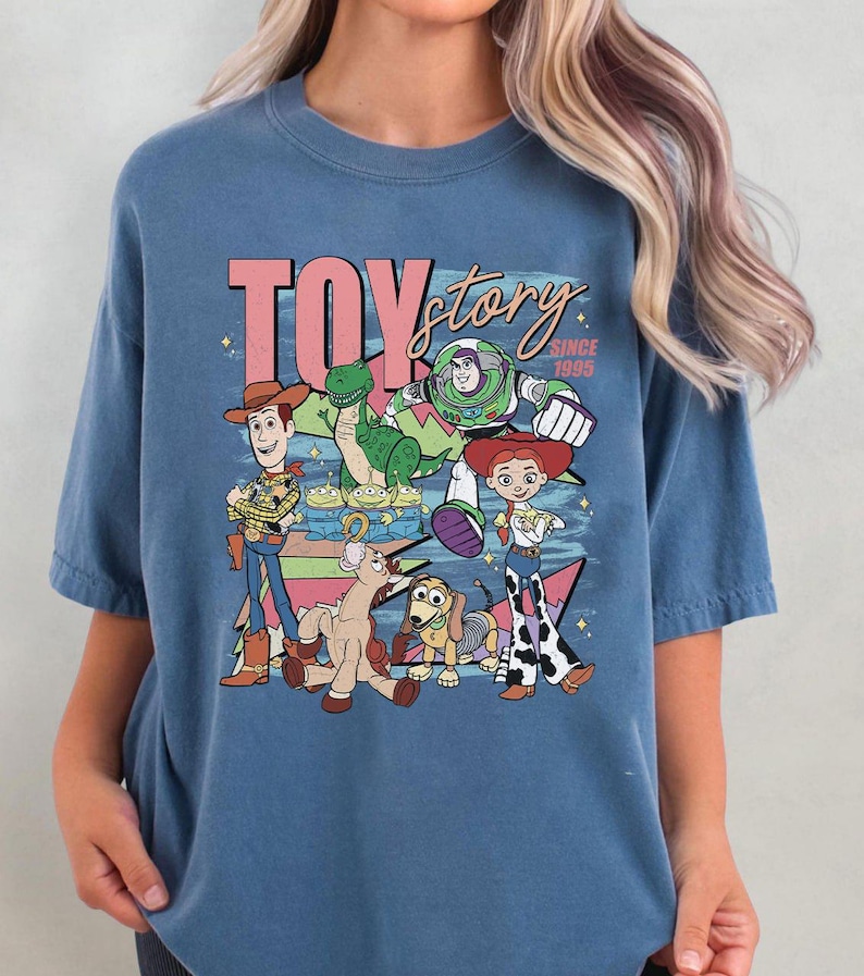 May include: Blue t-shirt featuring a graphic of characters from the animated film "Toy Story." The design includes the text "TOY story" in pink and white, along with various characters like Woody, Buzz Lightyear, and Jessie.