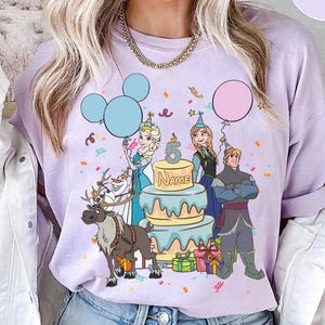 Puede incluir: Una sudadera lila con un gr&aacute;fico colorido de personajes de la pel&iacute;cula Frozen. El dise&ntilde;o incluye a Elsa, Anna, Kristoff, Olaf y Sven, junto con una tarta de cumplea&ntilde;os, globos y confeti. El n&uacute;mero 5 y la palabra "Name" tambi&eacute;n son visibles.