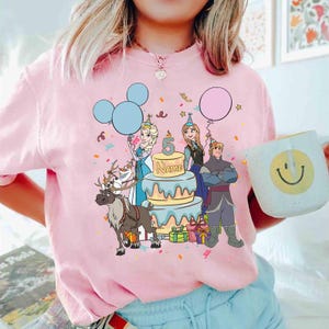 Puede incluir: Sudadera rosa de cuello redondo con un gr&aacute;fico colorido de personajes de Frozen, una tarta de cumplea&ntilde;os, globos y confeti. El n&uacute;mero 5 y la palabra "NAME" est&aacute;n en la tarta. Se sostiene una taza blanca con una carita sonriente.