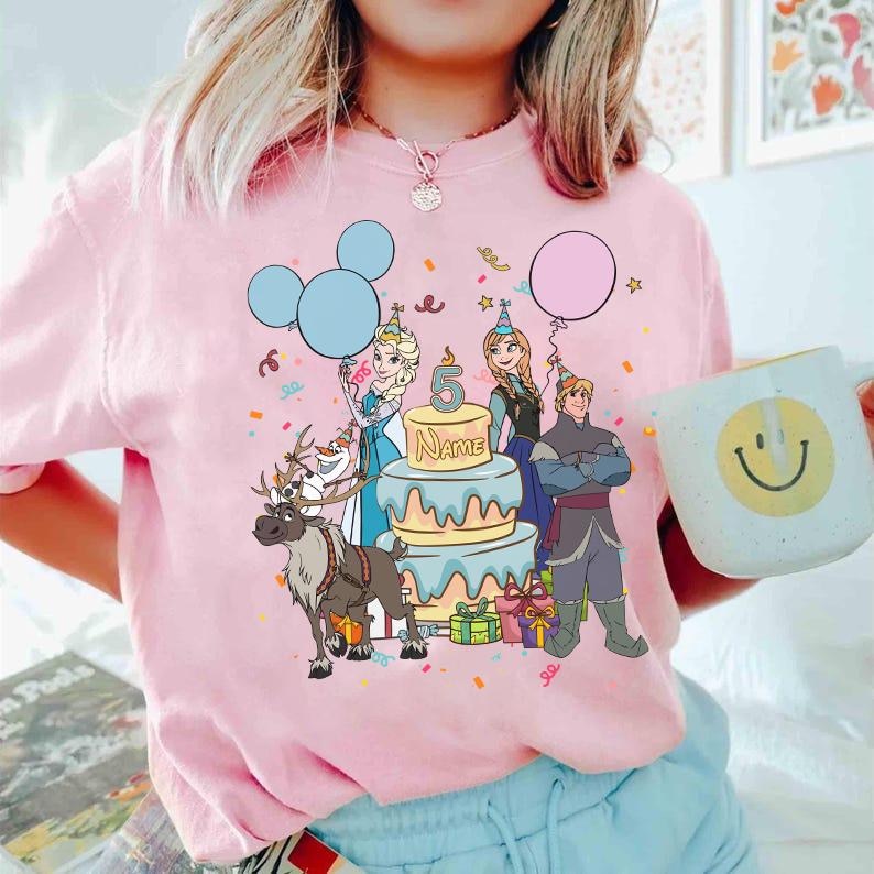 Puede incluir: Sudadera rosa de cuello redondo con un gr&aacute;fico colorido de personajes de Frozen, una tarta de cumplea&ntilde;os, globos y confeti. El n&uacute;mero 5 y la palabra "NAME" est&aacute;n en la tarta. Se sostiene una taza blanca con una carita sonriente.
