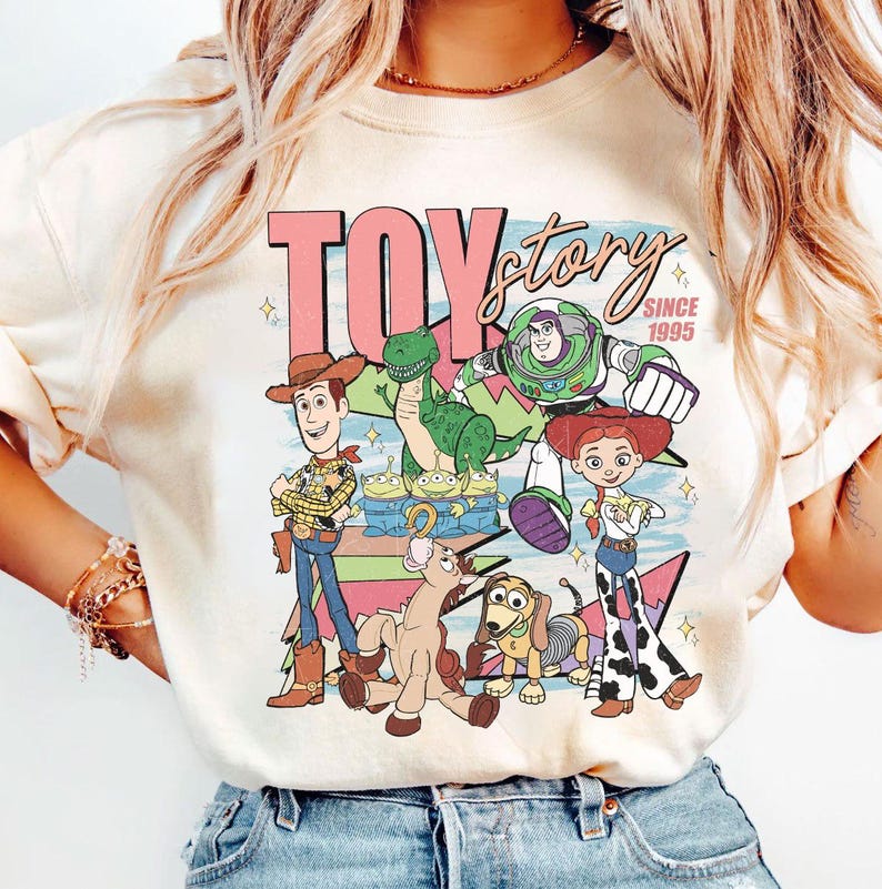 May include: Cream-colored t-shirt featuring a colorful graphic of Toy Story characters, including Woody, Buzz Lightyear, and Rex. The text "TOY STORY" is displayed in pink, with "SINCE 1995" below. A fun, nostalgic design.